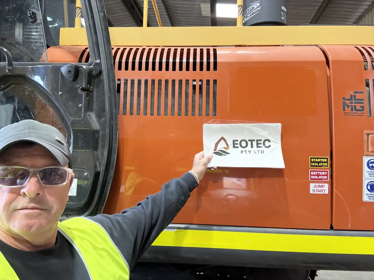 Tim Weir in high-visibility clothing beside an orange excavator, holding an EOTEC Pty Ltd sign.